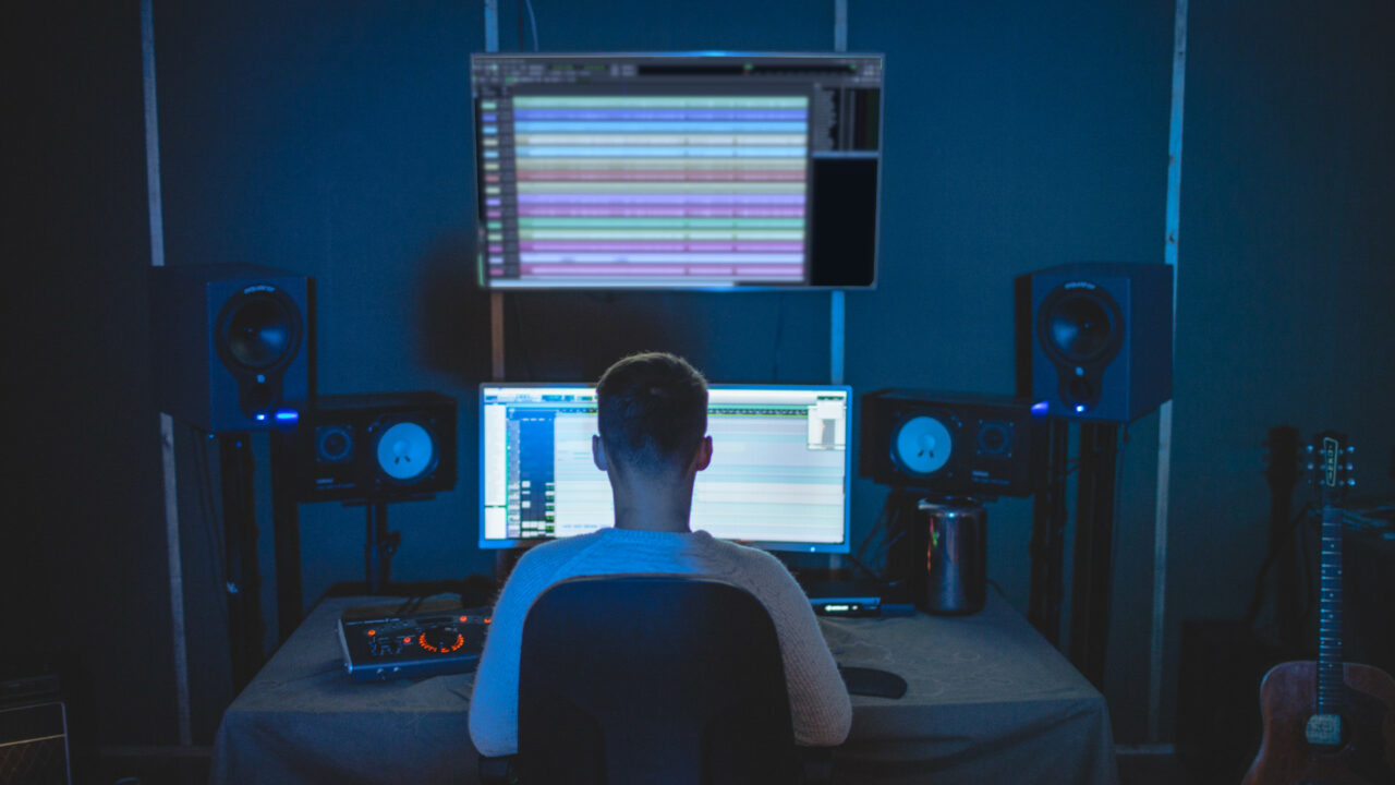 Mensch sitzt im Musikstudio vor zwei übereinander angeordneten Bildschirmen, daneben Lautsprecherboxen und eine Akustikgitarre